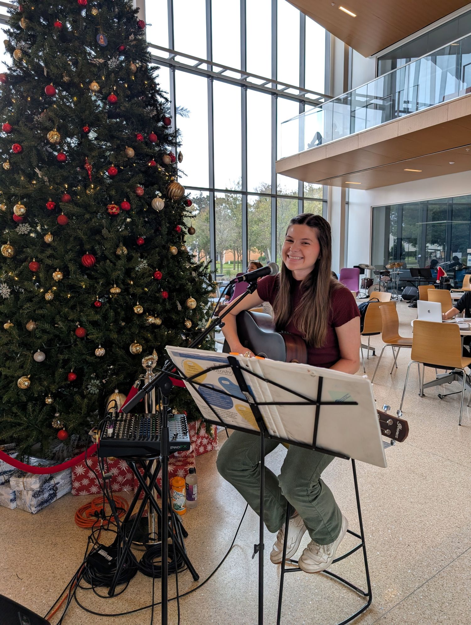 A woman sits on a stool playing a guitar beside a large decorated Christmas tree inside the Lemerand Student Center.
