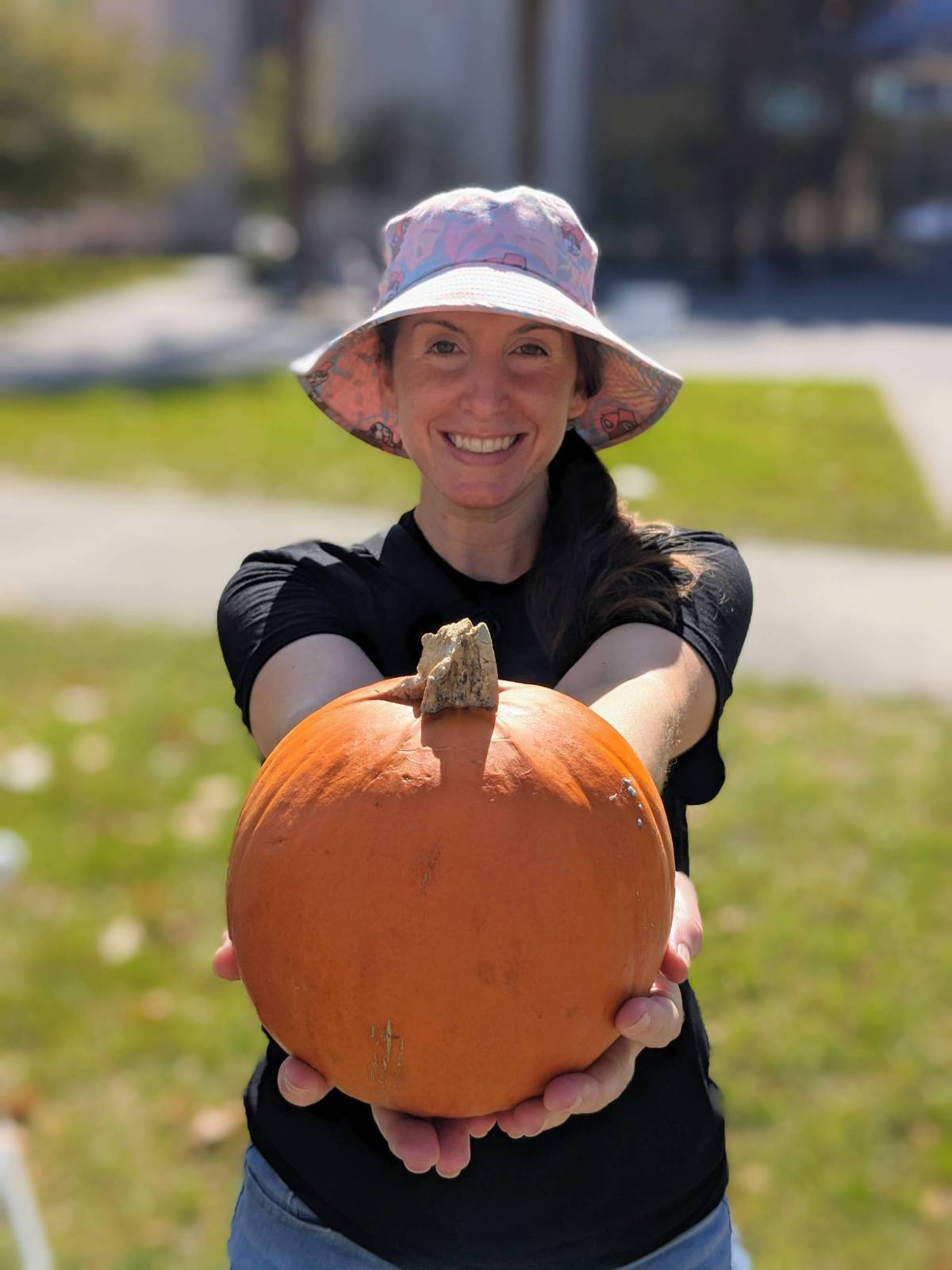 Female student with a floppy hat and a big smile holding a small pumkin with arms extended